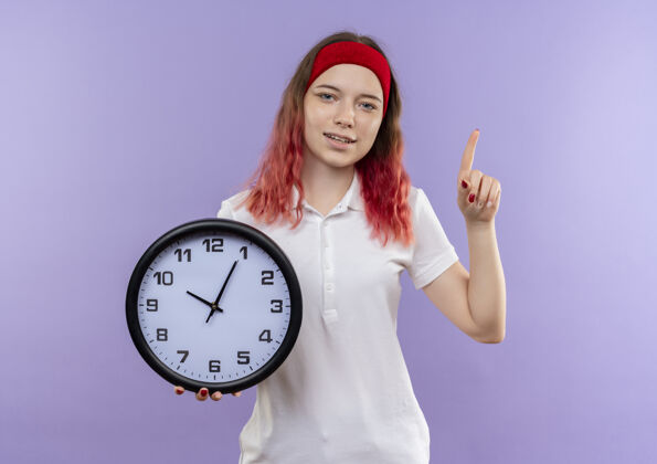 运动装年轻的运动型女人拿着挂钟 自信地指着紫色的墙壁指着女人看