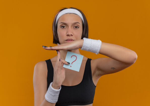 运动身穿运动服的年轻健身女 头戴头巾 手拿带问号的提醒纸 手拿严肃的脸 站在橙色的墙上 做着暂停的手势健康手人
