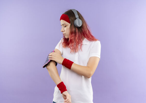 智能手机戴着头带 头戴耳机 站在紫色墙壁上触摸智能手机袖标的年轻运动女性公民女性站立