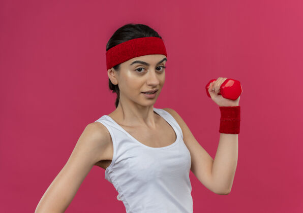手身穿运动服 头箍 举着哑铃的年轻健身女士站在粉红色的墙上 看起来很自信地做着运动健身姿势看