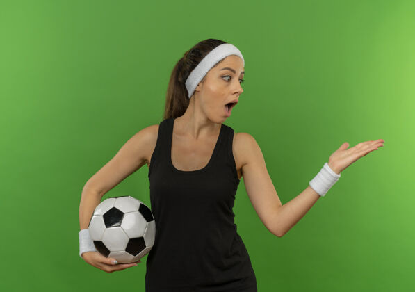 抱着身穿运动服的年轻健身女 头戴足球带 手举足球 站在绿色的墙上 脸上露出惊讶的神色足球女人头带