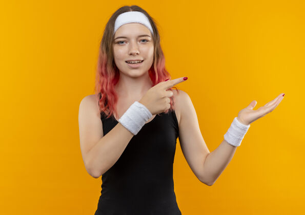 展示身着运动服的年轻健身女士微笑着用食指指着侧面 手臂站在橙色的墙上公动健康