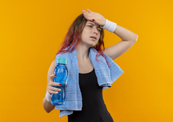 健身年轻的健身女孩穿着运动服 脖子上挂着毛巾 手里拿着一瓶看起来不舒服的水 站在橘色的墙上摸着她的头触摸人站