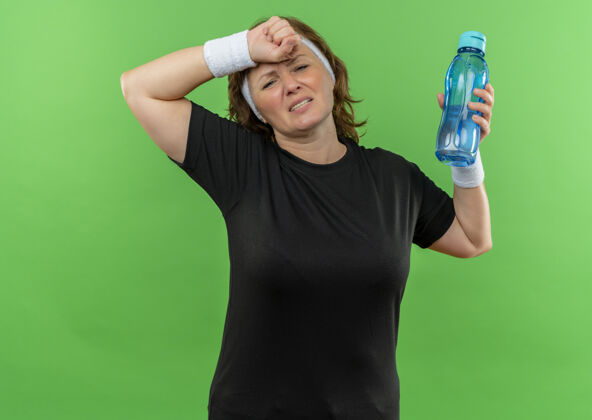运动中年运动女性 穿着黑色t恤 头上戴着头巾 手里拿着一瓶水 看起来很累 恼怒的表情抚摸着站在绿墙上的头女人表情运动员