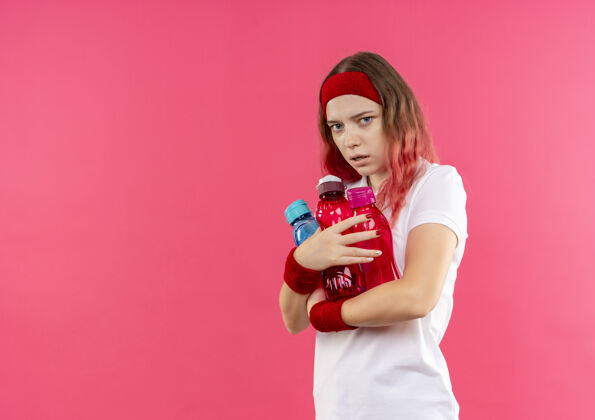 水戴着头巾的年轻运动女郎站在粉红色的墙上 抱着几瓶水 脸上带着恐惧的表情站运动运动装