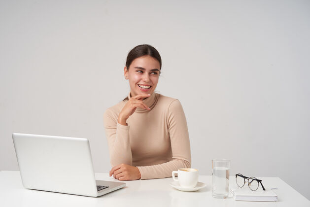 眼镜快乐的年轻漂亮的黑发女人微笑着 一边看着一边 把下巴靠在举起的手上 一边休息一边喝着咖啡 隔着白墙笔记本电脑爱情杯子
