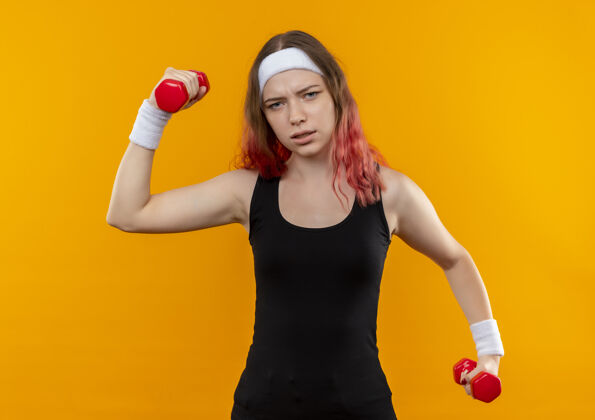 运动装穿着运动服的年轻健身女士手拿哑铃 站在橘色的墙上做运动 看起来很累手运动员看