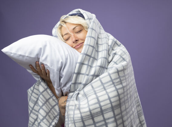 头生病的不健康的女人 短发 戴着暖和的帽子 裹着毯子 抱着枕头 把头靠在枕头上 闭着眼睛 在紫色的背景上微笑关闭包短裤