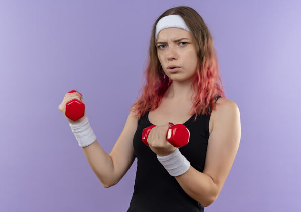 严肃穿着运动服的年轻健身女士站在紫色的墙上用哑铃做练习 脸严重拉伤姿势哑铃站立
