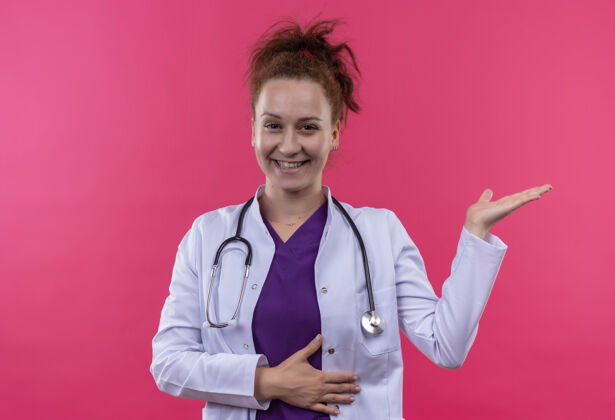 佩戴年轻的女医生身穿白色外套 手持听诊器 微笑着站在粉红色的墙上年轻人女人演示