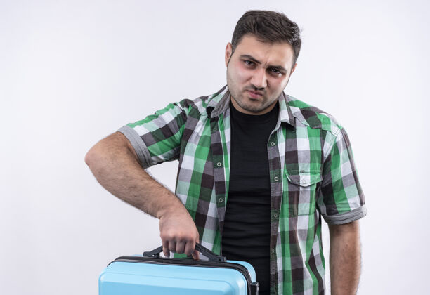 男人穿着格子衬衫的年轻旅行家手拿手提箱 脸上带着悲伤的表情站在白墙上检查脸抱着