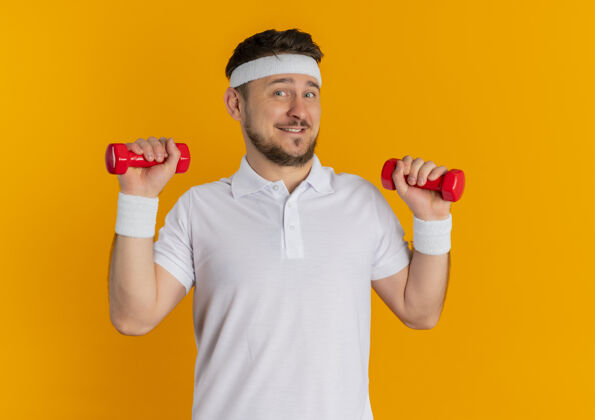 运动身穿白衬衫 头箍 举着哑铃的年轻健身男子站在橙色的墙上自信地微笑着站立教练运动装