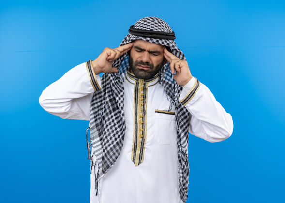 男人一位身着传统服装的阿拉伯男子站在蓝色的墙上摸着太阳穴 头痛得厉害强壮站立痛苦