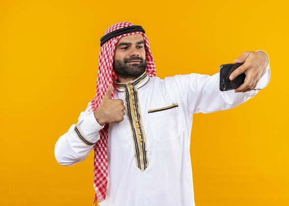 公民身着传统服装的阿拉伯商人站在橙色的墙上 用智能手机自拍 看上去很自信自拍看穿