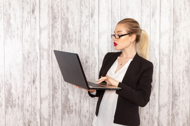 工作正面图穿着黑色夹克的年轻女商人在白色表面上使用她的笔记本电脑工作服严格女商务人士