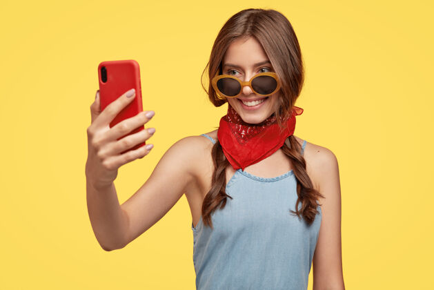 她自己穿着时髦的服装 戴着墨镜 手持红色手机 自拍肖像 微笑着 对着黄色的墙壁摆姿势年轻 科技和娱乐的概念黑发青少年电话