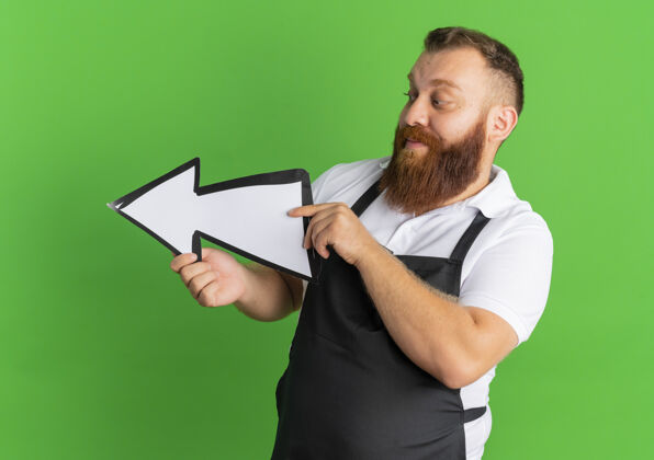 胡子围裙上的专业胡须理发师手举大箭头指向左边 站在绿色的墙上姿势站着拿着