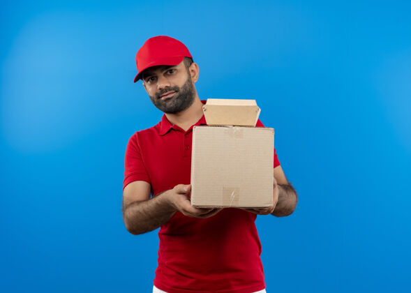 递送一个留着胡须的送货员 穿着红色制服 戴着帽子 手里拿着纸板箱 脸上带着微笑站在蓝色的墙上望着一边帽子站着制服