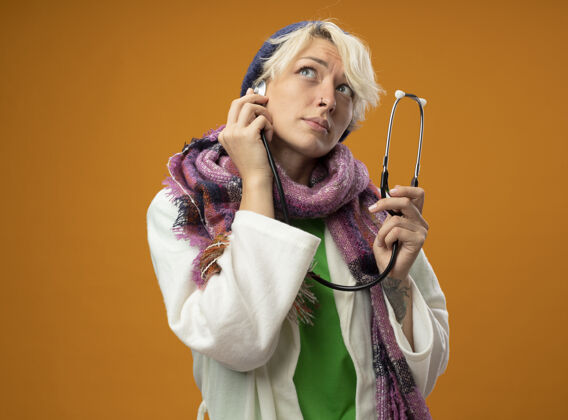 不健病的不健康的女人 短头发 戴着暖和的围巾 戴着听诊器 站在橙色的背景上困惑地抬头看帽子立场头发