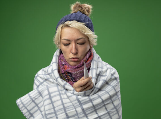 不健病的不健康的女人 短头发 戴着暖和的围巾和帽子 感觉不舒服 裹着毯子 拿着温度计站在绿色的背景上围巾头发毯子