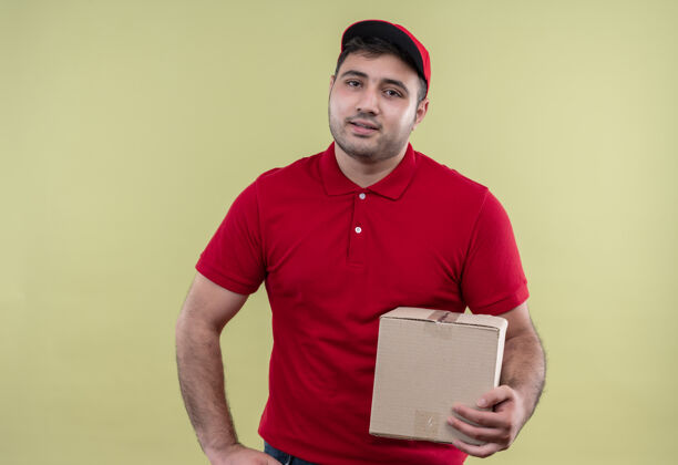 包装年轻的送货员穿着红色制服 戴着帽子拿着盒子 友好地站在绿色的墙上微笑着男人送货帽子