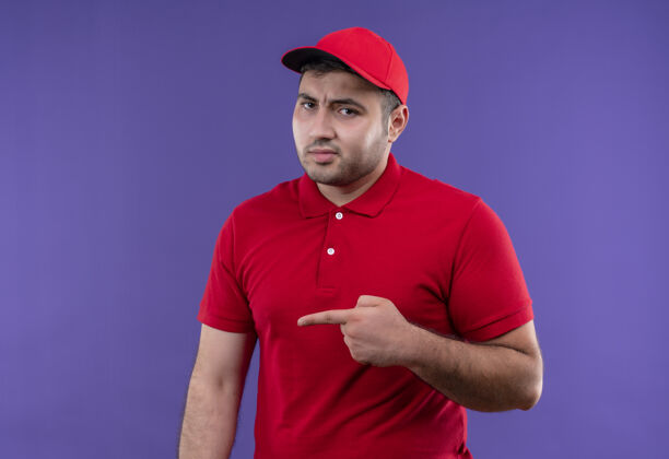侧线身穿红色制服 戴着帽子的年轻送货员站在紫色的墙壁上 手指指着侧面 不高兴地说指着姿势帽子