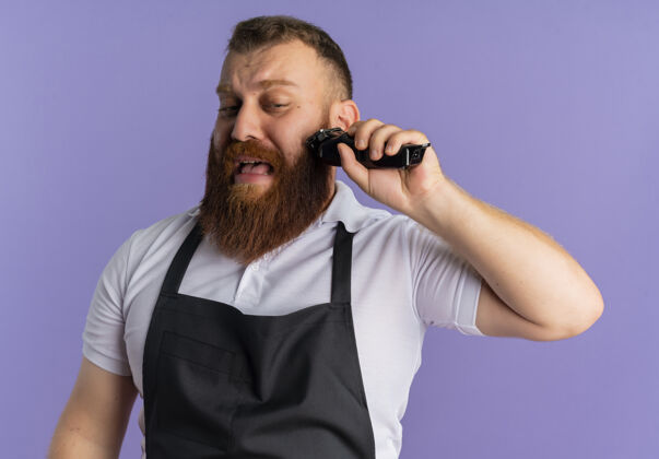 理发师专业的胡须理发师 围着围裙 用剃须刀剪胡子 站在紫色的墙上 脸上带着悲伤的表情表情专业胡须
