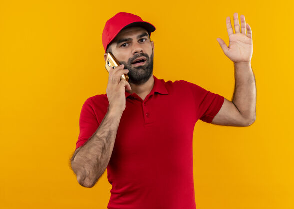 手机站在橘红色的墙上 一个留着胡须 身穿红色制服 戴着帽子的送货员正在讲手机 看上去很困惑 很焦虑说话站着电话