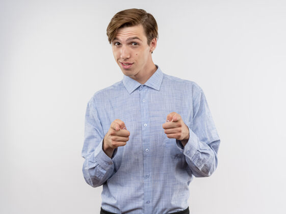 立场身着蓝色衬衫的年轻人用食指指着镜头 站在白色背景下 微笑着 自信 快乐 积极微笑指向积极