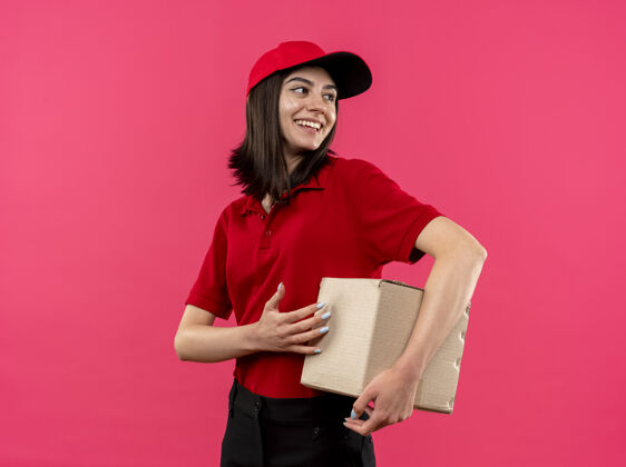 马球穿着红色马球衫和帽子的年轻送货女孩拿着盒子包 站在粉色背景下 脸上带着幸福的微笑看着一边脸包装立场