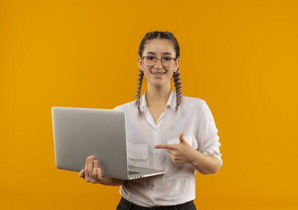 指着戴着眼镜 梳着辫子 穿着白衬衫的年轻女学生拿着笔记本电脑 手指指着笔记本电脑 自信地微笑着站在橙色的墙上辫子人手指