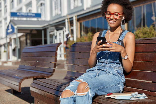 街道照片中快乐的黑人妇女手持手机 输入短信 使用耳机 听音乐 穿着破烂的工作服 在户外做模特 享受播放列表现代科技 在线交流钢笔手机无线