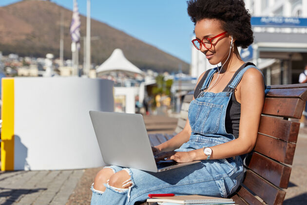 高兴一个黑人发型的黑皮肤女人的侧拍 在笔记本电脑上看视频 膝盖上拿着设备 坐在新鲜空气中的长凳上 在户外学习 当代电子小玩意