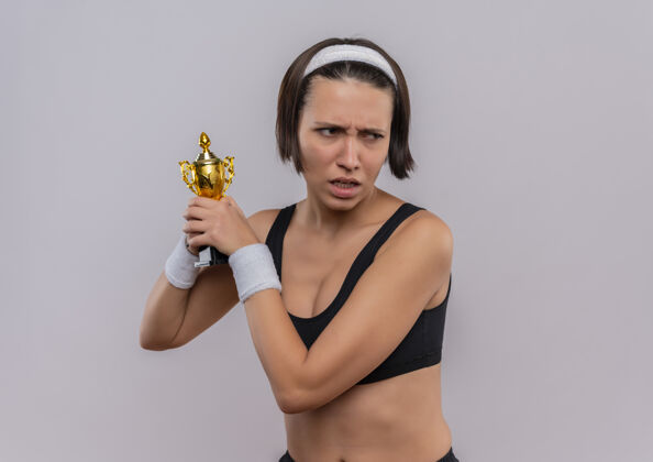 运动装身穿运动服的年轻健身女士手持她的奖杯 眉头紧锁地站在白色的墙上 望向一边人奖杯市民