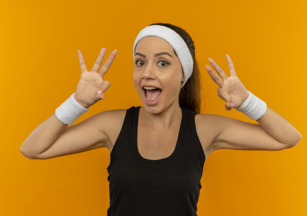 表演身穿运动服的年轻健身女 头戴花边 微笑着 乐观地站在橙色的墙上 显示“ok”的标志人市动