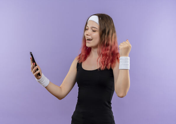 姿势身穿运动服的年轻健身女士手持智能手机 握紧拳头 高兴地站在紫色的墙壁上走了出来抱着拳头女人