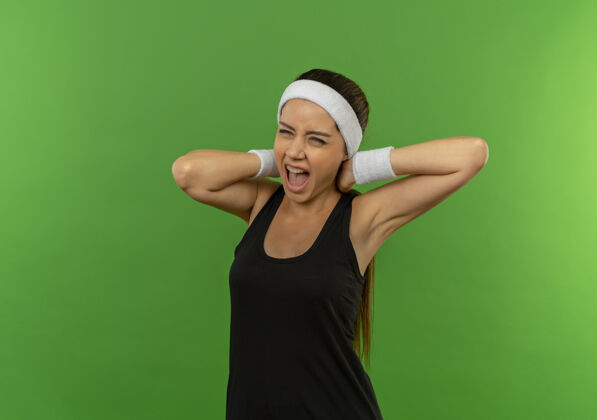 表情穿着运动服的年轻健身女士站在绿色的墙壁上 用恼怒的表情抚摸着脖子大声喊叫健身人市民