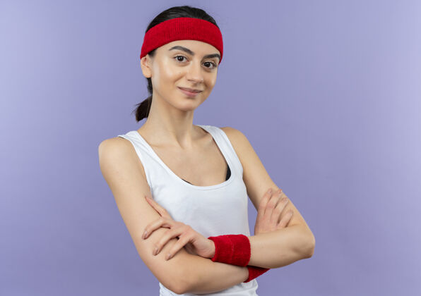交叉身穿运动服的年轻健身女 头戴头带 自信地微笑着 双手交叉放在胸前 站在紫色的墙上女人运动装自信