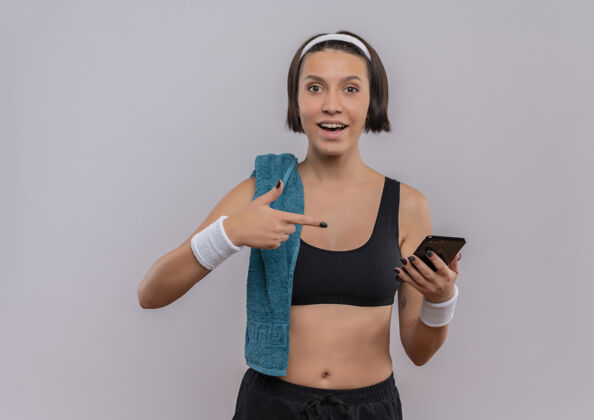 运动员身穿运动服的年轻健身女士 肩上挎着毛巾 站在白墙上 用手指着智能手机微笑着移动姿势运动