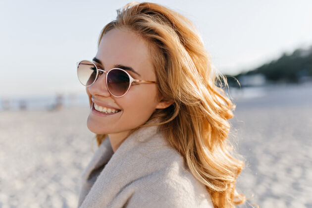 米色毛衣海滩上穿着米色羊绒衫 戴着太阳镜 一头卷曲的盲发的积极女人的特写照片寒冷年轻海边
