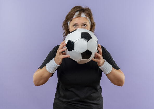 女人身穿黑色t恤 头箍 手持足球 表情自信 严肃的中年运动女性站在蓝色的墙上运动运动装年龄