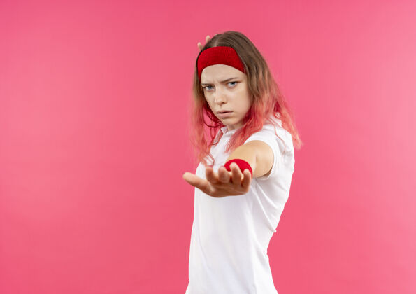 运动戴着头巾的年轻运动女郎站在粉红色的墙上 用一张严肃的脸做着停车标志脸头带看