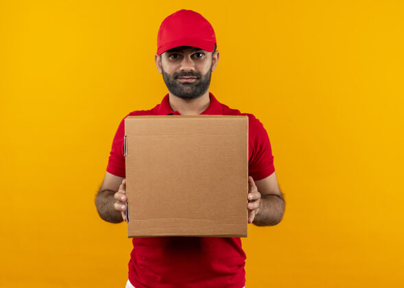 男人满脸胡须的送货员身着红色制服 戴着帽子 手拿盒子 表情自信严肃地站在橙色的墙上帽子胡子送货