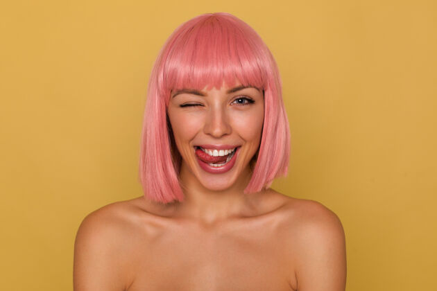 前锁风骚的年轻漂亮快乐的粉红色头发的女性 鲍勃的发型 快乐地看着镜头 伸出舌头 站在芥末墙的一边高兴地向镜头眨眼肩膀年轻封闭