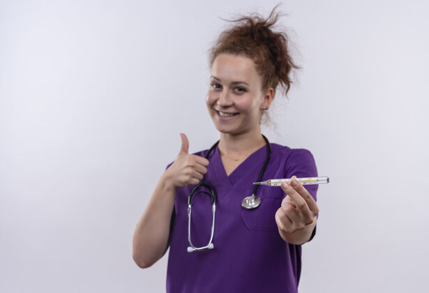 抱着年轻的女医生穿着医疗制服手持听诊器微笑着竖起大拇指站在白色的墙上展示制服戴着