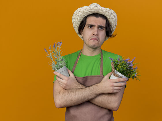 站着不快乐的年轻园丁 围着围裙 戴着帽子 手里拿着瘦弱的植物 站在橙色背景下 带着悲伤的表情看着摄像机围裙园丁不开心