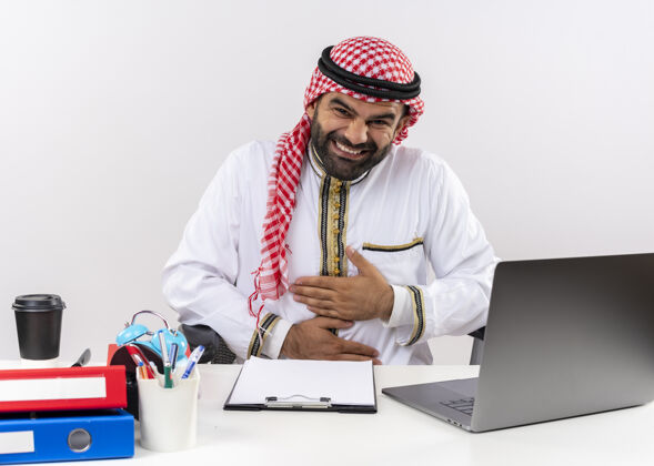 穿身着传统服装的阿拉伯商人坐在桌旁 拿着笔记本电脑笑着摸着肚子在办公室工作肚子桌子坐