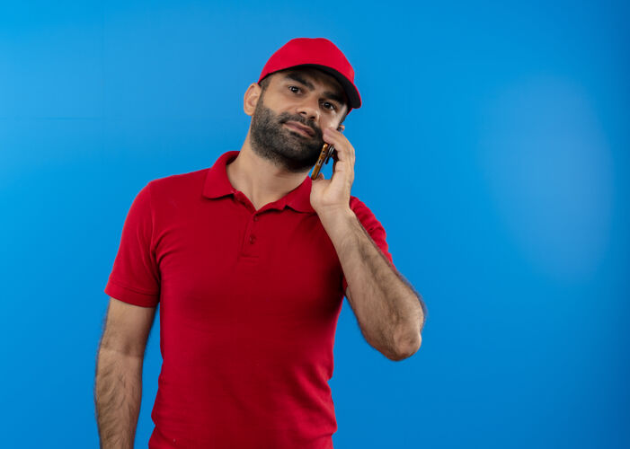 站有胡子的送货员穿着红色制服 戴着帽子 站在蓝色的墙上讲着手机 看上去很自信帽子信心看