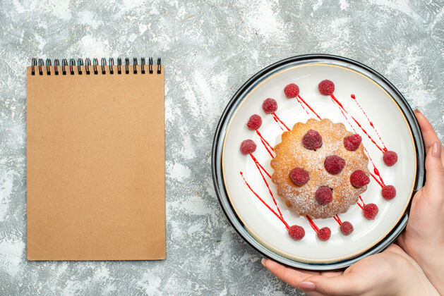 Oval俯视图白色椭圆形盘子上的浆果蛋糕在灰色表面上的女性手记事本化妆粉Berrycake