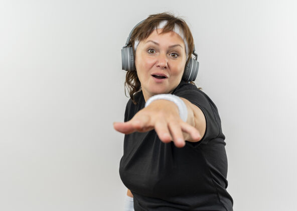 指着中年运动女性 身穿黑色t恤 头戴头带 头戴耳机 手臂站在白墙上 看上去很惊讶惊喜耳机手臂